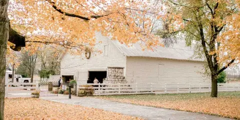 Exterior view of a white historic barn with a stone chimney, surrounded by white fencing and autumn trees.