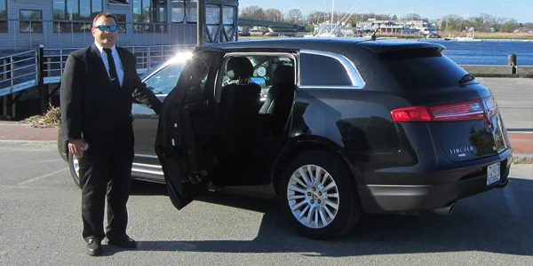 A professional chauffeur in a black suit stands beside a black Lincoln MKT with the rear door open, parked near a waterfront.