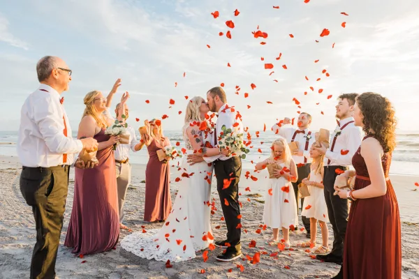 A bride and groom kissing on a sandy beach during a ceremony while guests throw red rose petals into the air.