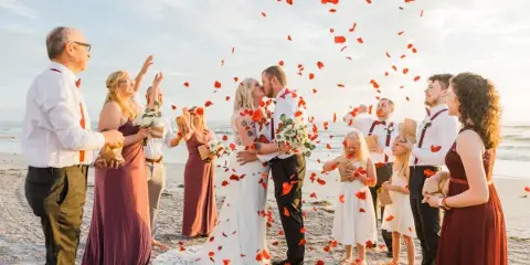 A bride and groom kissing on a sandy beach during a ceremony while guests throw red rose petals into the air.