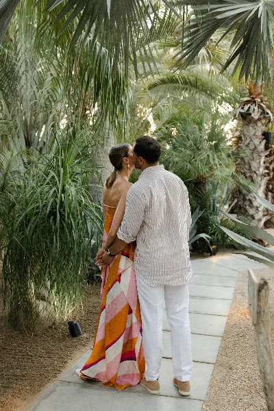 A couple standing in a tropical garden setting, the woman wearing a vibrant, patterned dress and the man in a striped shirt, holding hands.