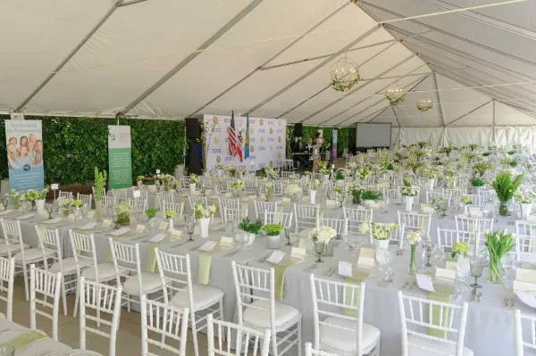 White event tent interior with multiple round tables dressed in white linens, white Chiavari chairs, floral centerpieces, and hanging chandeliers.