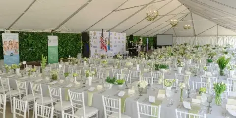 White event tent interior with multiple round tables dressed in white linens, white Chiavari chairs, floral centerpieces, and hanging chandeliers.