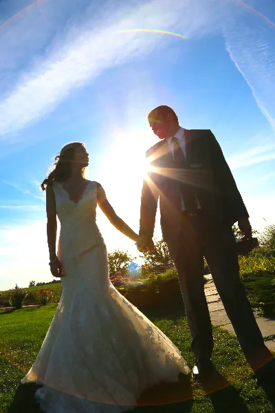 A bride and groom holding hands and looking at each other in a grassy field with the sun flaring behind them.