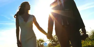 A bride and groom holding hands and looking at each other in a grassy field with the sun flaring behind them.