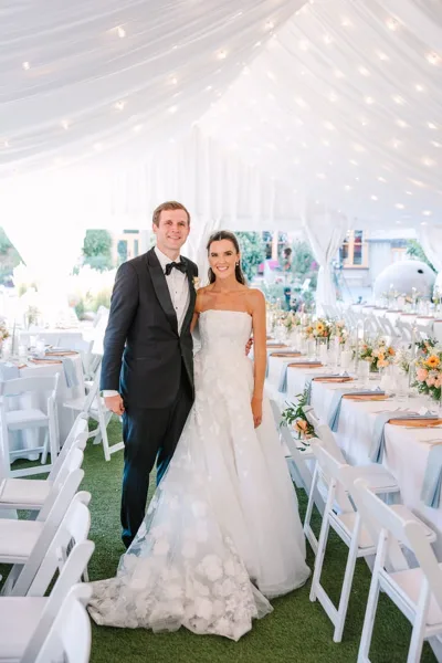 A bride and groom standing together at a wedding reception with the bride wearing a white floral gown and styled hair.