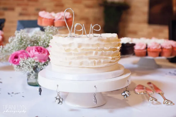 A white ruffled wedding cake with a wire 'Love' topper displayed on a white cake stand, surrounded by pink flowers and cupcakes.