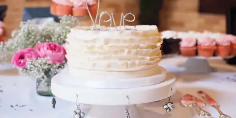 A white ruffled wedding cake with a wire 'Love' topper displayed on a white cake stand, surrounded by pink flowers and cupcakes.