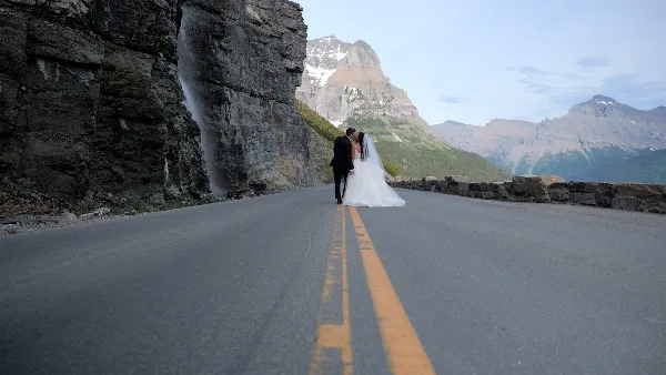 A bride and groom kissing on a paved road with a waterfall and mountains in the background.