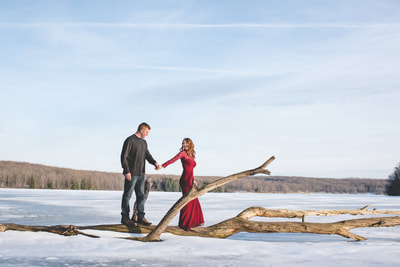 A couple posing together on a fallen tree in a snowy field under a clear sky.