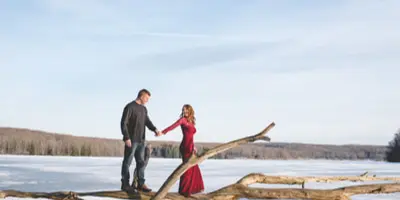 A couple posing together on a fallen tree in a snowy field under a clear sky.