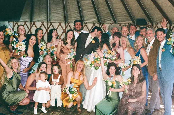 A candid, documentary-style group photograph of a wedding party and guests celebrating together under a tent.