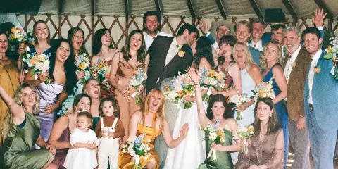 A candid, documentary-style group photograph of a wedding party and guests celebrating together under a tent.