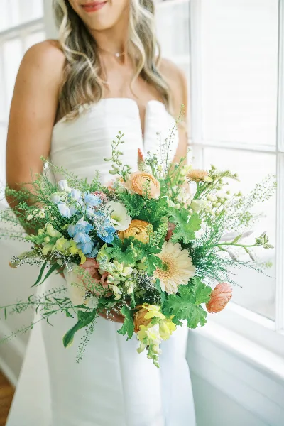 A bride in a white dress holding a large, colorful floral bouquet with blue, yellow, and peach flowers and greenery.
