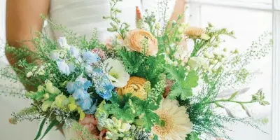 A bride in a white dress holding a large, colorful floral bouquet with blue, yellow, and peach flowers and greenery.