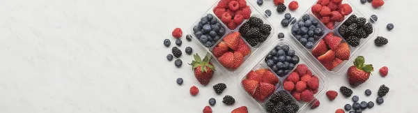 Overhead view of clear plastic containers filled with strawberries, blueberries, raspberries, and blackberries on a white marble surface.