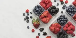 Overhead view of clear plastic containers filled with strawberries, blueberries, raspberries, and blackberries on a white marble surface.