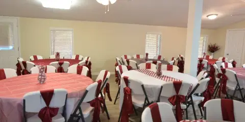 Interior view of an event venue with round tables set with red and white tablecloths and chairs decorated with red sashes.