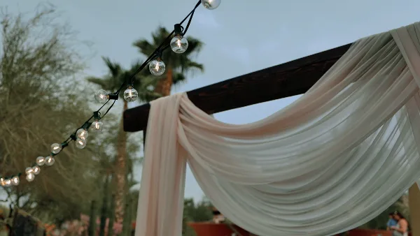 A wedding arch draped with white fabric and string lights against a backdrop of palm trees and a twilight sky.