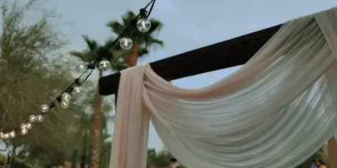 A wedding arch draped with white fabric and string lights against a backdrop of palm trees and a twilight sky.