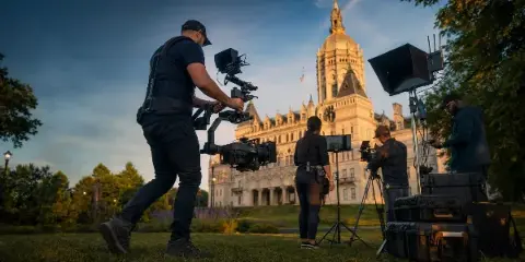 Video production crew operating gimbal-mounted cinema cameras, tripods, and lighting equipment on a lawn in front of a large domed state capitol building.