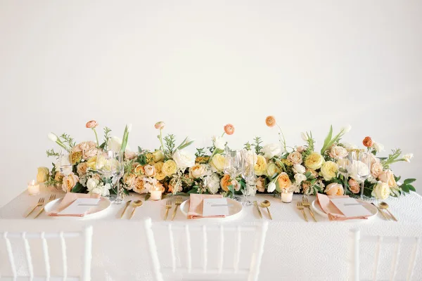 A professionally styled wedding table featuring a long floral centerpiece with yellow and white roses, gold cutlery, and white Chiavari chairs.