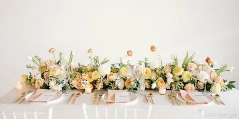 A professionally styled wedding table featuring a long floral centerpiece with yellow and white roses, gold cutlery, and white Chiavari chairs.