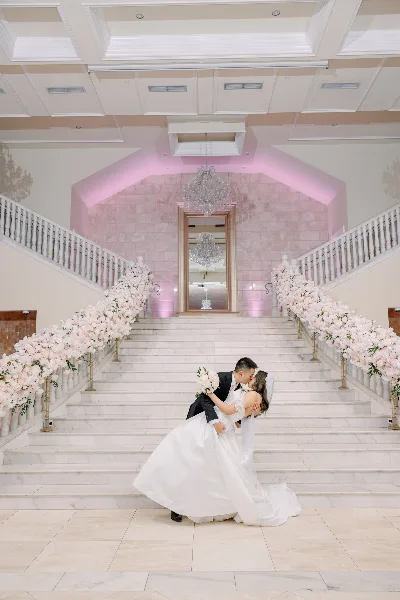A bride and groom sharing a kiss on a grand marble staircase decorated with floral arrangements and chandeliers.