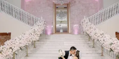 A bride and groom sharing a kiss on a grand marble staircase decorated with floral arrangements and chandeliers.