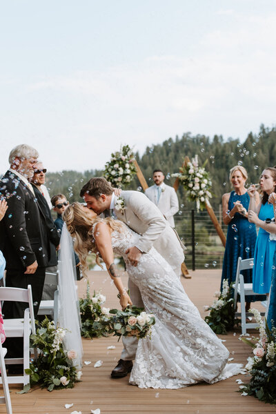 A bride and groom share a kiss during an outdoor wedding ceremony on a wooden deck, surrounded by guests and floral decorations.