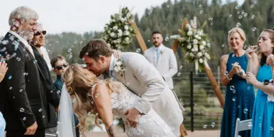 A bride and groom share a kiss during an outdoor wedding ceremony on a wooden deck, surrounded by guests and floral decorations.