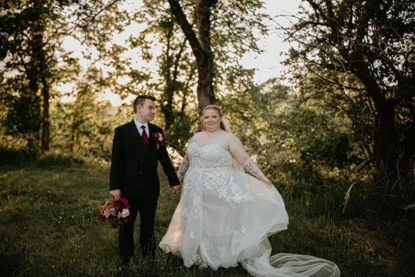 A bride and groom holding hands in a grassy field with trees in the background.