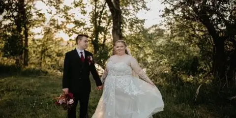 A bride and groom holding hands in a grassy field with trees in the background.