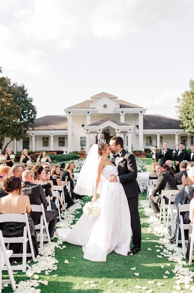 A bride and groom kissing at the end of a wedding aisle with seated guests and a white venue building in the background.