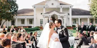 A bride and groom kissing at the end of a wedding aisle with seated guests and a white venue building in the background.