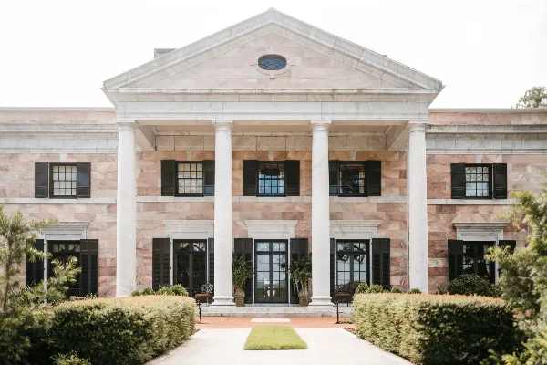 Exterior view of a large, historic stone mansion with a white columned portico and manicured hedges.