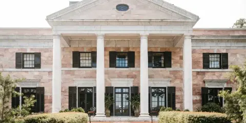 Exterior view of a large, historic stone mansion with a white columned portico and manicured hedges.