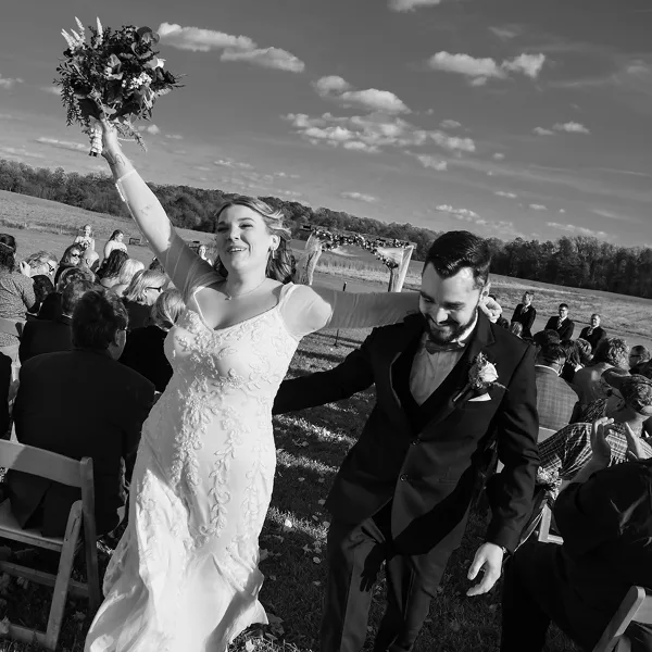 A black and white photograph of a bride and groom during an outdoor wedding ceremony, with the bride raising her bouquet and the groom smiling.