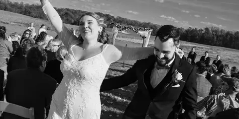 A black and white photograph of a bride and groom during an outdoor wedding ceremony, with the bride raising her bouquet and the groom smiling.