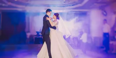 A bride and groom dancing at a wedding reception under purple stage lighting and smoke effects.