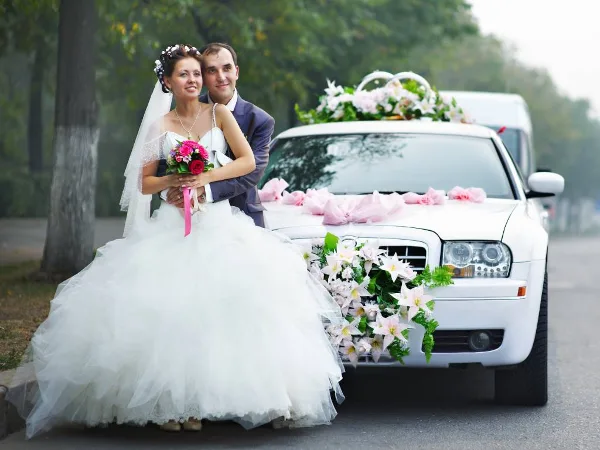 Bride and groom posing in front of a white wedding car decorated with flowers