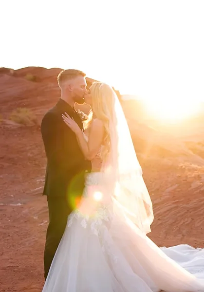 A bride and groom embracing in a romantic pose against a backdrop of red rock formations during a sunset.