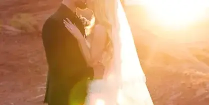 A bride and groom embracing in a romantic pose against a backdrop of red rock formations during a sunset.