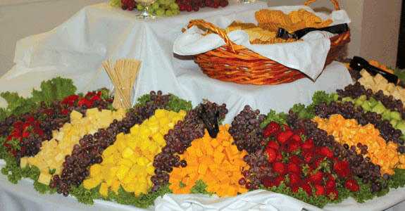 A catering table set with a large fruit and cheese display, including grapes, strawberries, and melon, alongside a basket of bread.