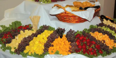 A catering table set with a large fruit and cheese display, including grapes, strawberries, and melon, alongside a basket of bread.