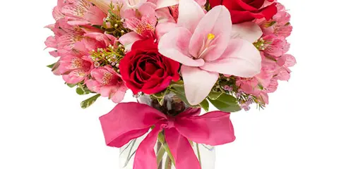 A hand-arranged floral bouquet containing red roses, pink lilies, and alstroemeria in a clear glass vase with a pink ribbon.