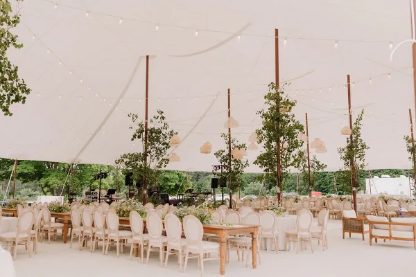 A large white tent interior set up for an event with rows of wooden tables, cream-colored chairs, and hanging string lights.