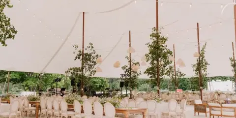 A large white tent interior set up for an event with rows of wooden tables, cream-colored chairs, and hanging string lights.