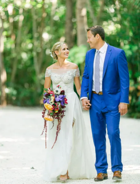 A bride in a white lace gown and a groom in a blue suit walking hand-in-hand on a path in a wooded area.