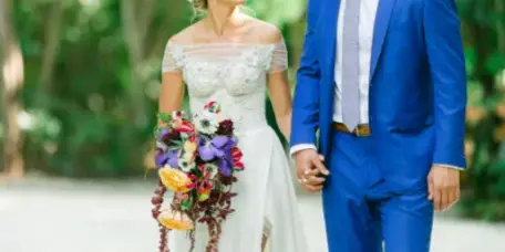 A bride in a white lace gown and a groom in a blue suit walking hand-in-hand on a path in a wooded area.